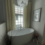 A modern bathroom features a white freestanding tub, a large window with beige curtains, a potted plant, a small stool, and a framed artwork on the wall.